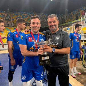 Two men pose with a large silver trophy after a win, surrounded by teammates in blue uniforms in an indoor arena.