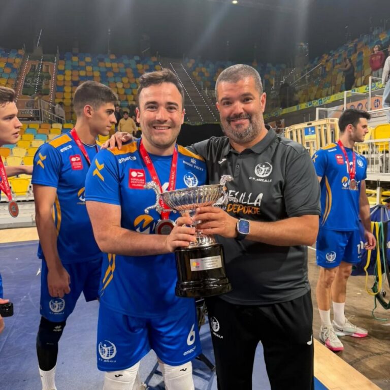 Two men pose with a large silver trophy after a win, surrounded by teammates in blue uniforms in an indoor arena.