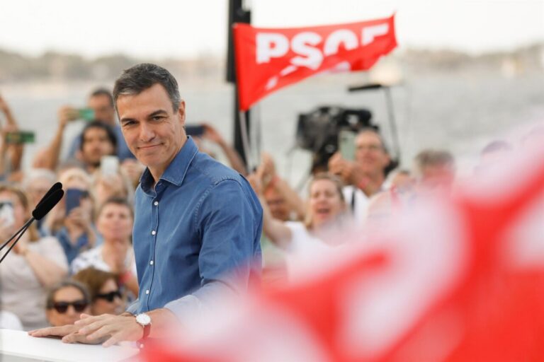 Man in blue shirt speaking at a podium during a political rally, with a PSOE banner and cheering crowd in the background