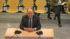 Elder man in a dark suit sits at a wooden podium with two microphones, speaking in a conference room.