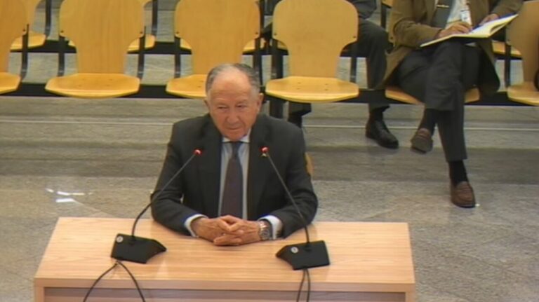 Elder man in a dark suit sits at a wooden podium with two microphones, speaking in a conference room.