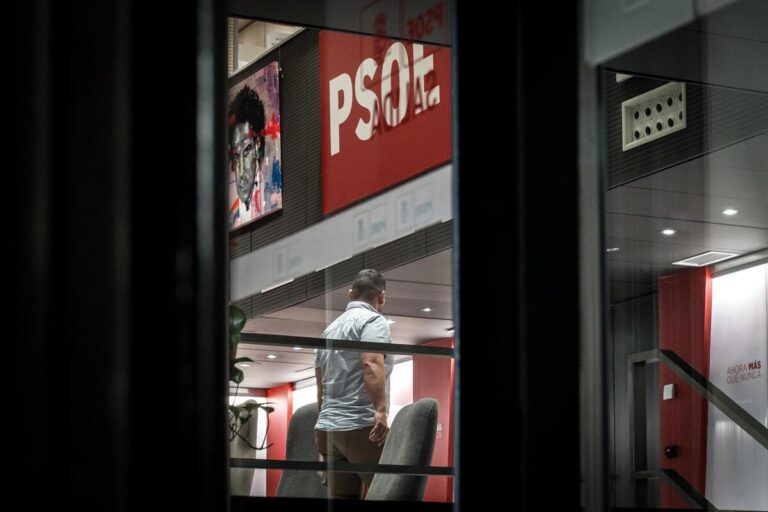 Man in a light shirt stands inside a modern office with red branding and large posters on the wall, viewed through a window pane.