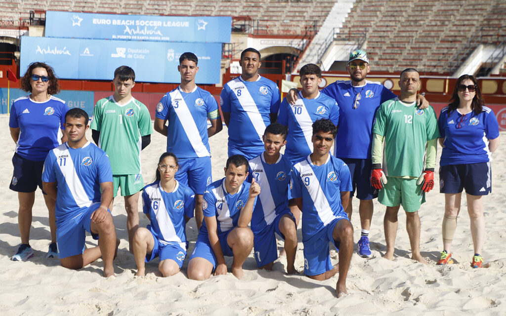 Beach soccer team posing for a group photo on the sand with blue and white uniforms and a goalkeeper in green, stadium banners in the background.