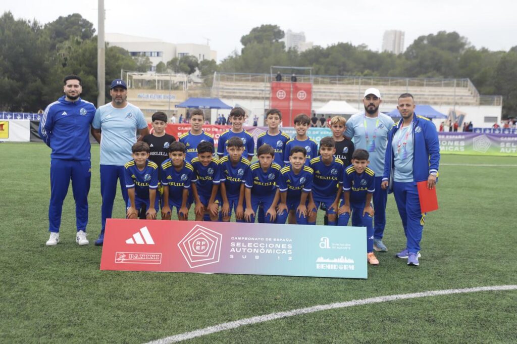 Youth soccer team in blue uniforms poses for a group photo on a grass field, with coaches standing behind them and a large banner in front.