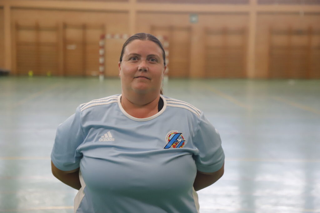 Woman in a light blue Adidas sports jersey stands with hands behind her back in an indoor gymnasium.