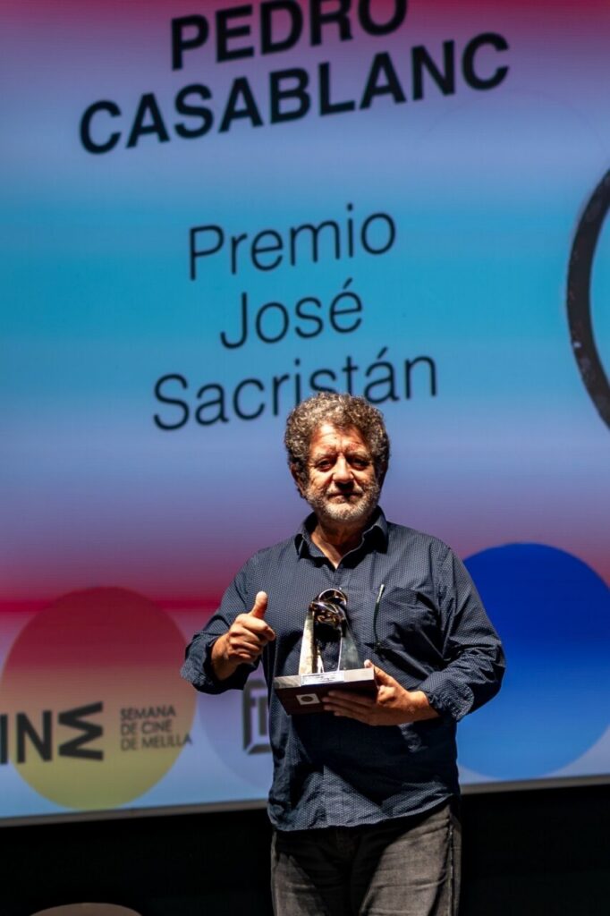 Man on stage holding a trophy and giving a thumbs‑up, with a backdrop reading 'Premio José Sacristán' behind him.