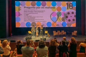 Three speakers stand center stage with microphones, colorful banner in background reading 'Pregunta por mí mañana' and 18 on the right, audience applauding.
