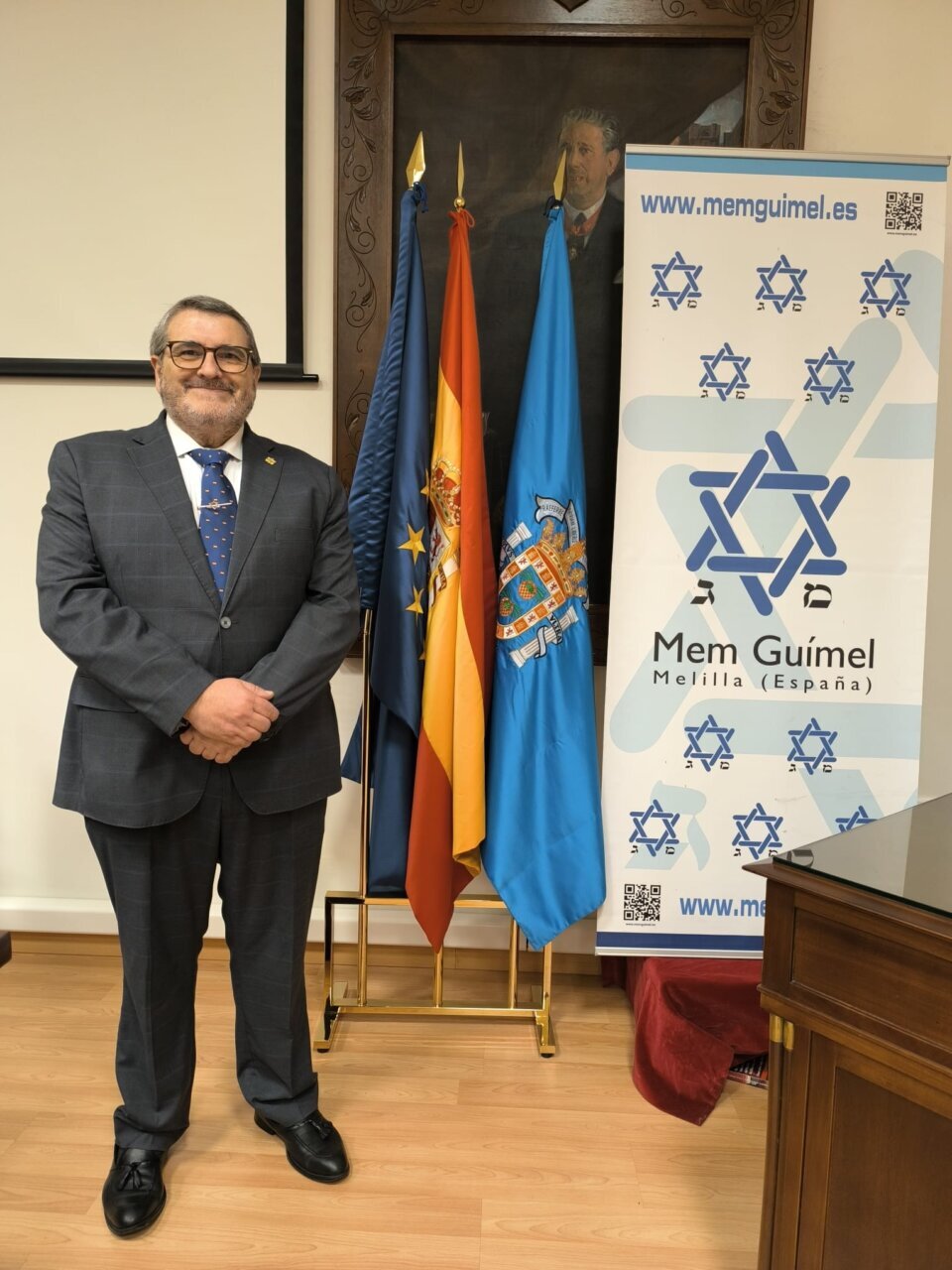 Hombre de pie junto a banderas y un banner de Mem Guímel
