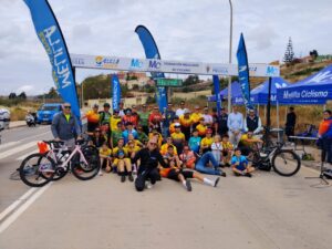 Large group of cyclists and supporters pose for a group photo at a race finish line with banners and blue tents in the background, bikes in the foreground.