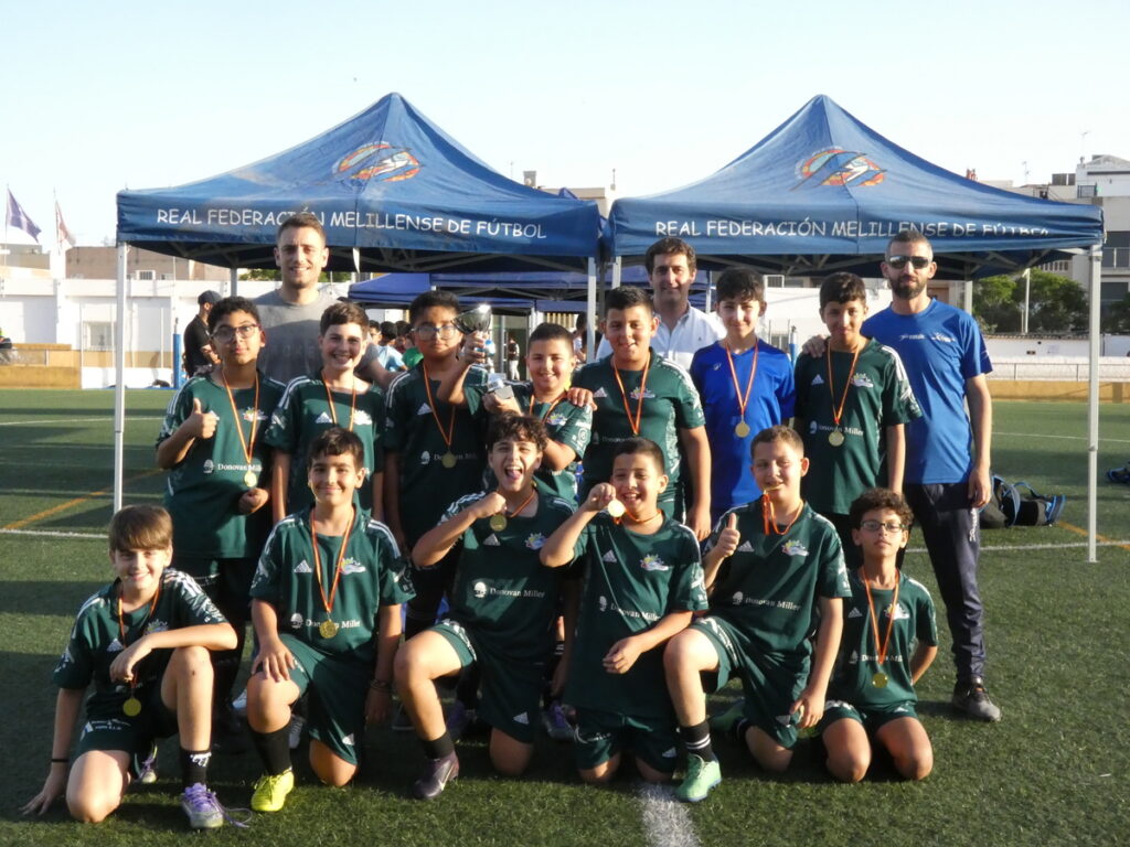 Youth soccer team posing on a field after a tournament, wearing green uniforms and medals with coaches in back under blue tents labeled 'Real Federación Melillense de Fútbol'.