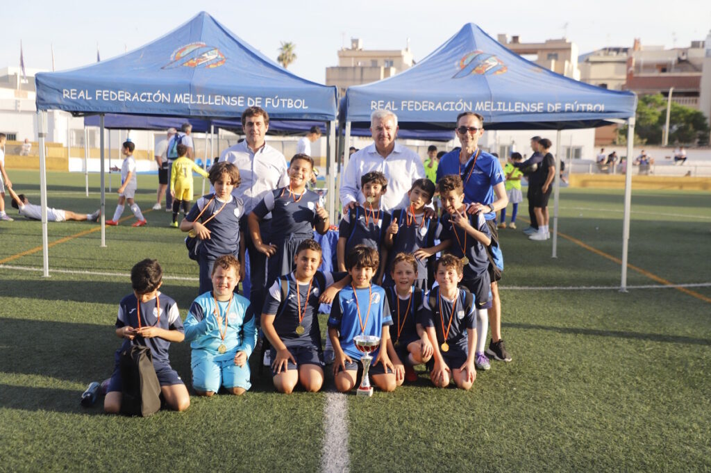 Youth soccer team posing for a group photo with coaches, wearing medals, and a trophy on a soccer field under blue tents that read 'Real Federación Melillense de Fútbol'.