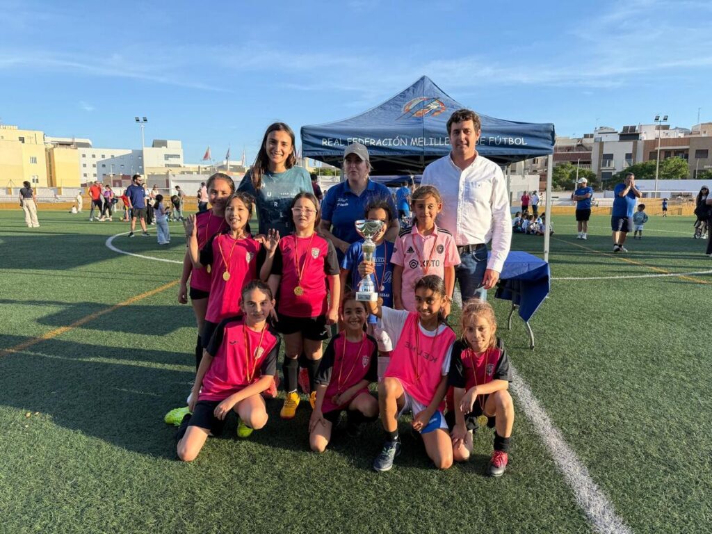 Girls' youth soccer team posing with a trophy on a sunny field, wearing pink jerseys with coaches nearby.