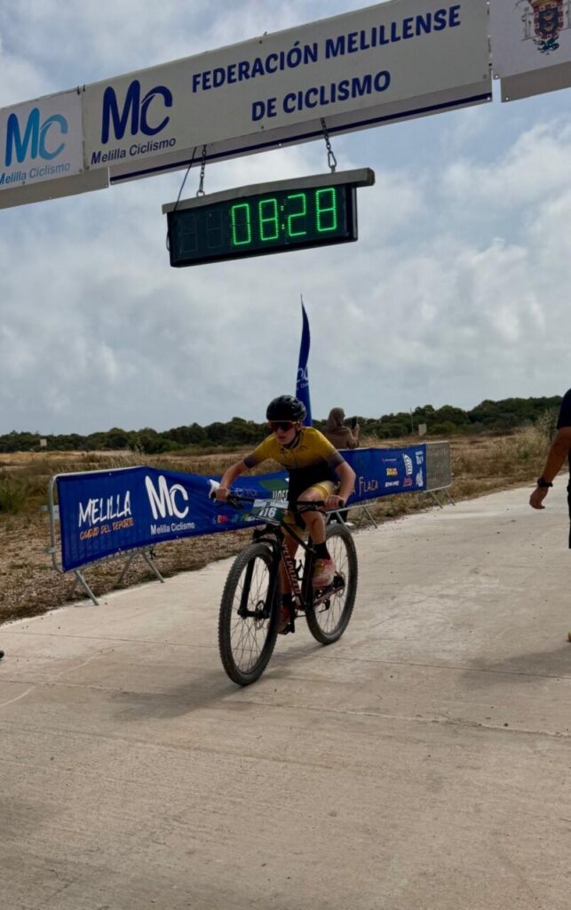 Cyclist in a yellow kit crosses the finish line under Melilla Ciclismo banners; clock shows 08:28