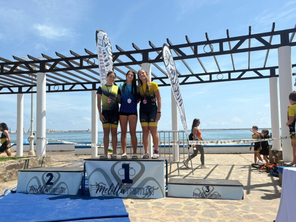Three young female triathletes stand on a winners' podium by the seaside, wearing medals against a bright blue sky at Melilla Triatlón.
