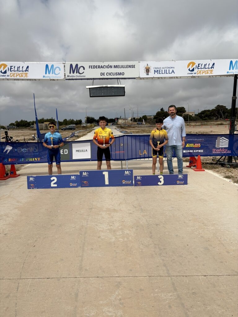 Three young cyclists stand on a winners' podium holding trophies, with an official beside them after a race.
