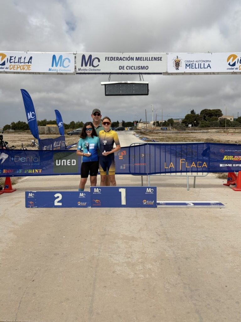 Two young cyclists stand on the winners' podium (1st and 2nd place), each holding a trophy, with a man behind them at a cycling finish line.