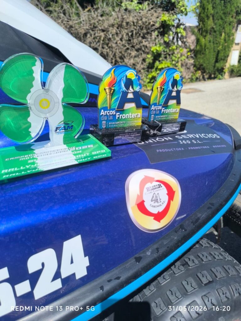 Three trophies displayed on a blue car hood, including two tall blue plaques and a green flower-shaped award, with a sticker and tire nearby in a sunny outdoor setting.