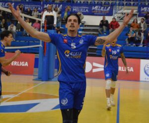 Volleyball player in blue jersey #5 raises both arms in celebration on an indoor court; teammate and spectators cheer in the background.
