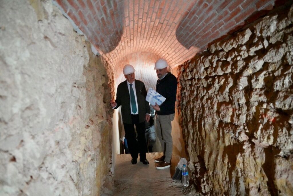 Two men wearing hard hats study blueprints inside a brick arch tunnel during a site visit.