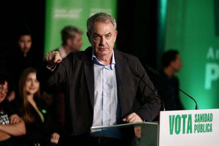 Man in a dark suit points toward the audience from a podium, with a green campaign backdrop and a sign reading 'VOTA SANIDAD PÚBLICA'