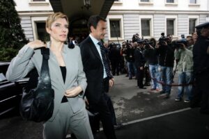Businesswoman in a gray suit walking away from a crowd of photographers outside a light-colored building, carrying a large black bag.