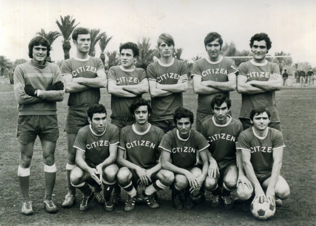 Eleven soccer players in matching ‘CITIZEN’ jerseys pose for a team photo on a field, some standing, others kneeling with a ball nearby.