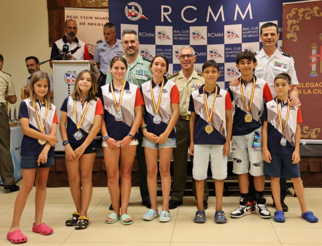 Eight children in matching navy, white, and red shirts with medals pose on a stage with officials behind them at a maritime club event.