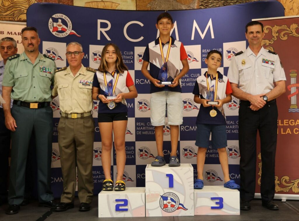 Three young medalists stand on a winners' podium holding trophies and medals, flanked by uniformed officials at a maritime club ceremony.