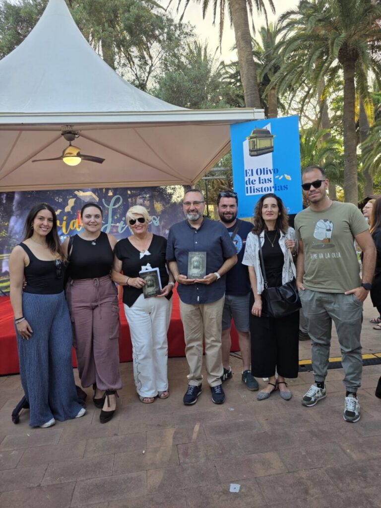 Eight adults posing together outdoors at a book event under a tent, some holding books.