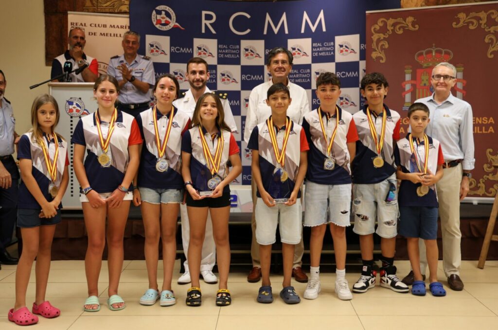Group of children wearing matching uniforms with gold medals posing for a daytime awards ceremony, adults and banners behind them.