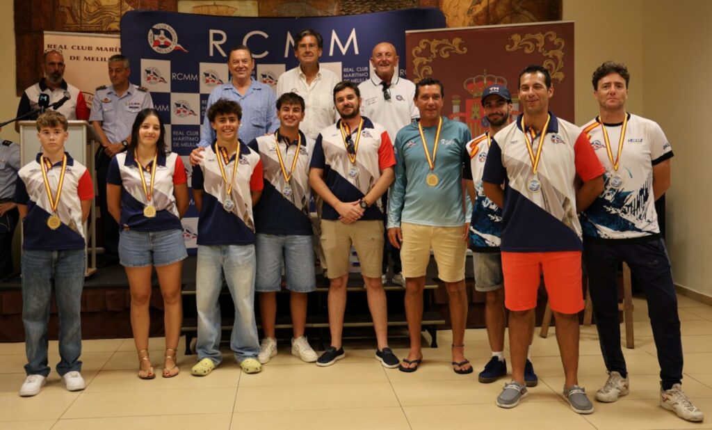 Group of athletes standing in a row on a stage, wearing matching white, blue, and red uniforms with medals, posing for a photo in front of a RCMM backdrop and event banners.