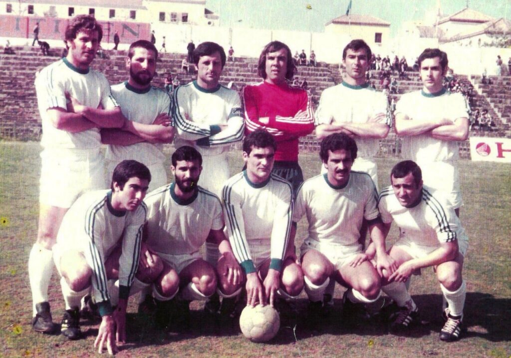 Vintage soccer team posing on a field with a ball in front; players wear white uniforms with green trim, red goalie shirt in back row.