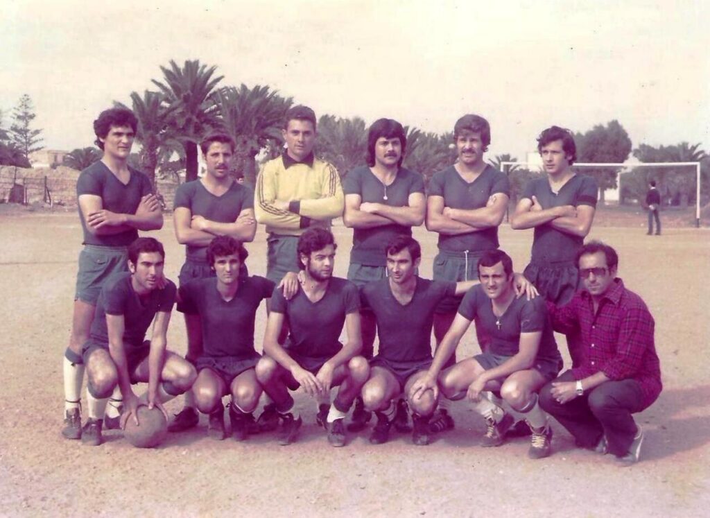 Soccer team photo: ten men in green uniforms posing on a dusty field with palm trees and a goal in the background.