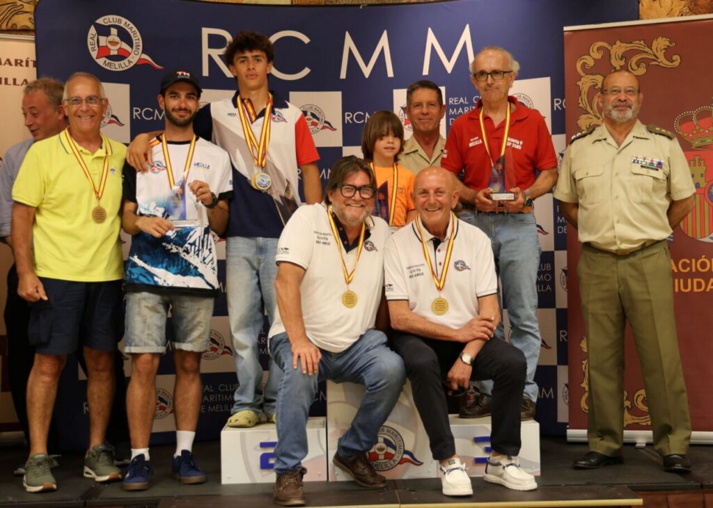 Group of medalists and officials posing on a podium after a maritime club awards ceremony, blue backdrop with logos behind them.