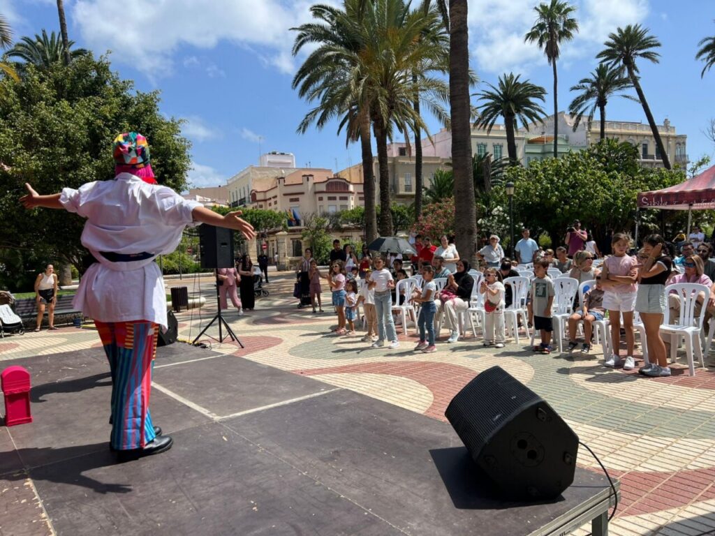 Clown on stage with arms outstretched performing for children seated in white chairs in a sunny palm-filled plaza.