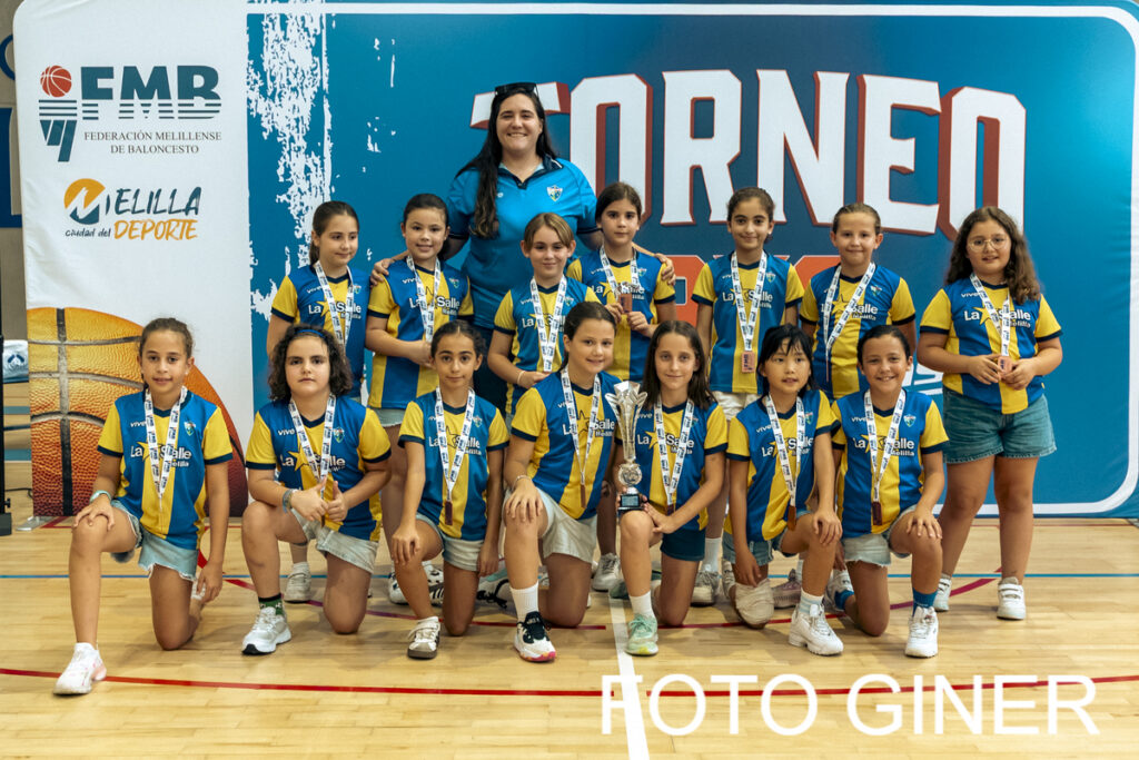 Girls' basketball team posing with medals and a trophy in a gym after a tournament, coach in blue behind them on a blue backdrop reading 'TORNEO'.