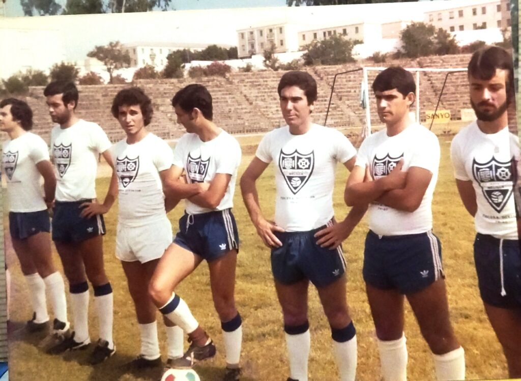 Vintage soccer team photo: seven players in white shirts with crest, blue shorts, and knee-high socks posing on a grass field with Bleacher stands in the background.