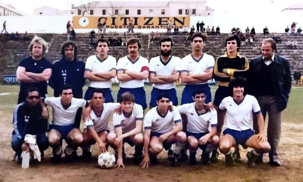 A soccer team poses for a group photo on the field, players in white shirts and blue shorts with coaches standing at the sides.