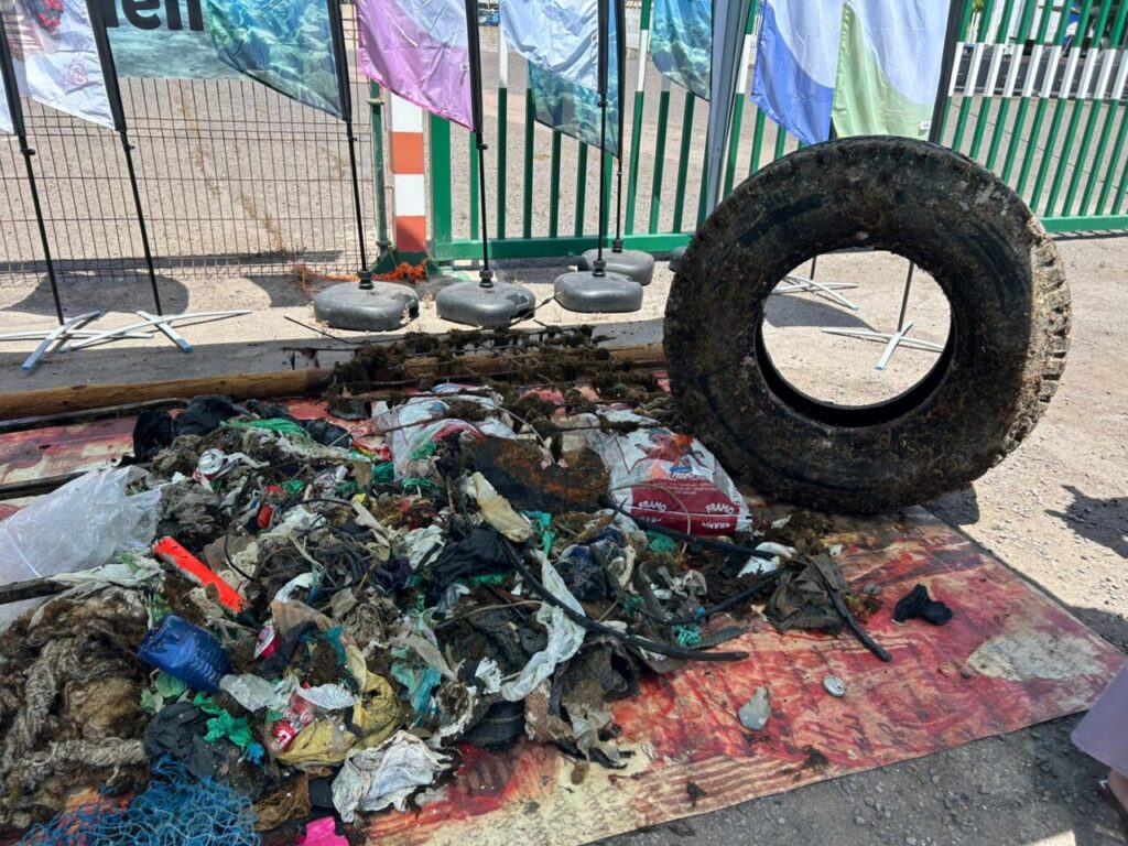 Pile of dirty trash and debris on a red tarp, with a large used tire propped nearby and banners in the background.