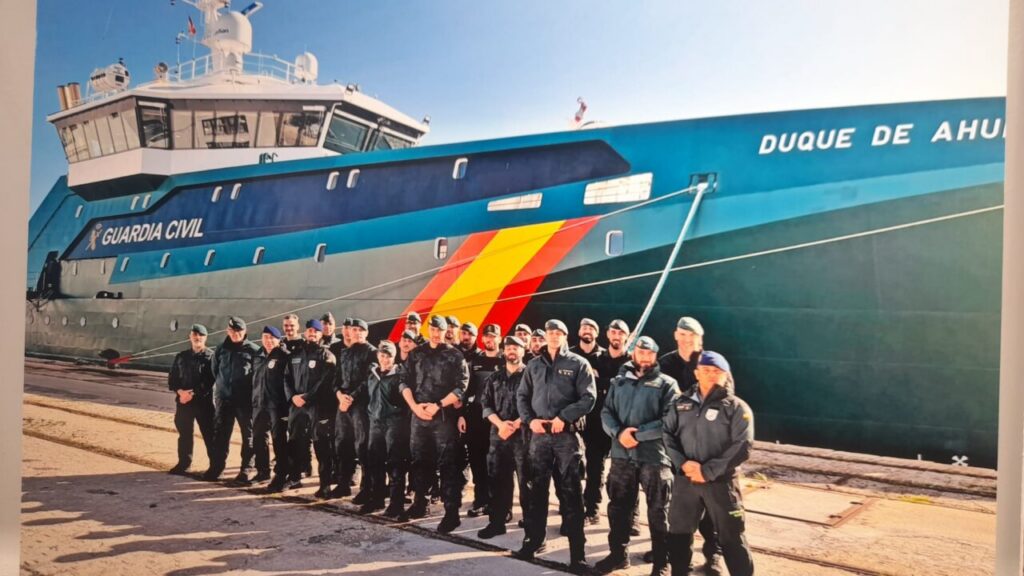 Group of Guardia Civil officers in uniform posing in front of a large blue patrol vessel docked at the quay, with Spanish flag colors on the hull.