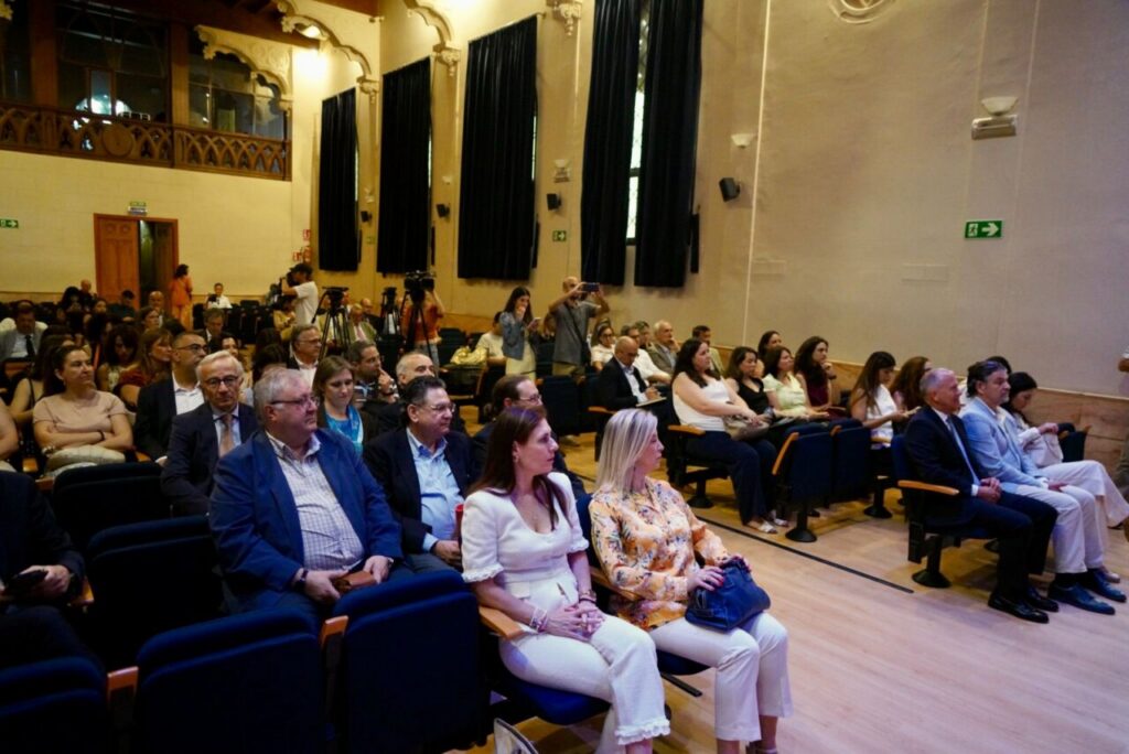 Audience seated in a formal auditorium watching a event, with several people in business attire and a few taking photos in the back.