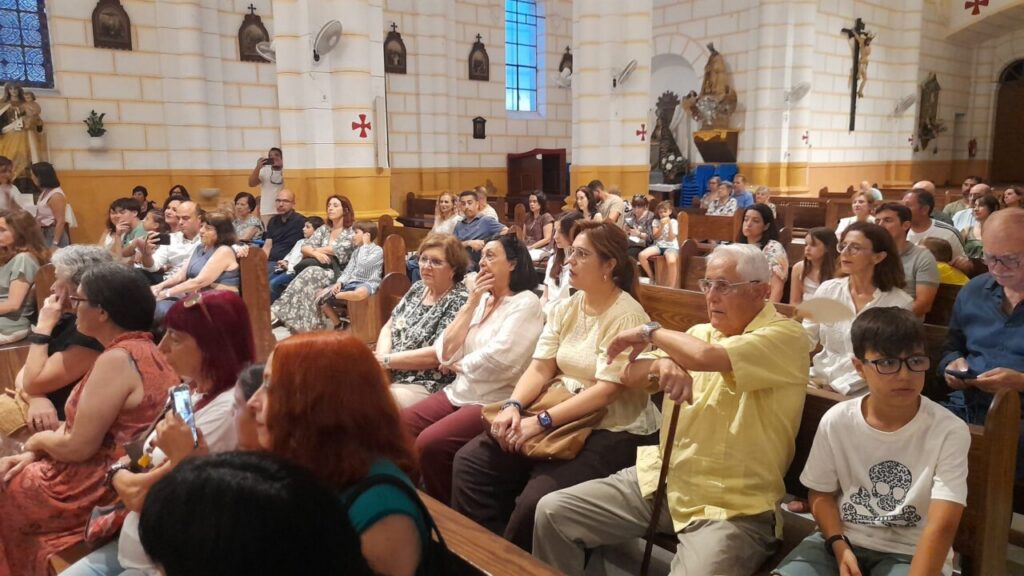 People seated in a church nave watching a ceremony, with columns and religious statues in the background.