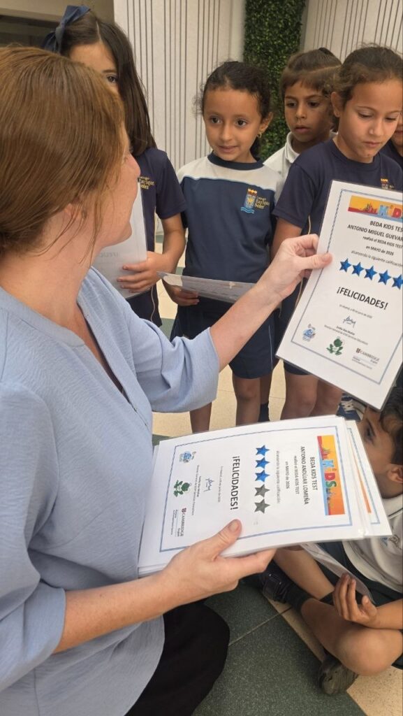 Teacher hands out certificates to a group of schoolchildren in uniform, standing in a hallway/courtyard.