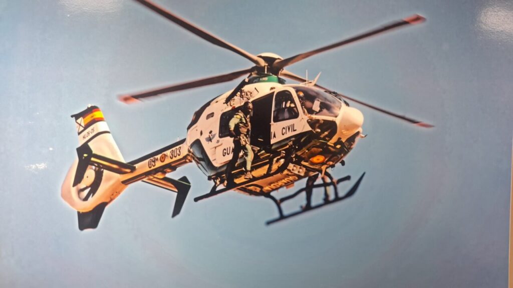 White rescue/civil helicopter in mid-air with a crew member climbing into the open side door against a blue sky.