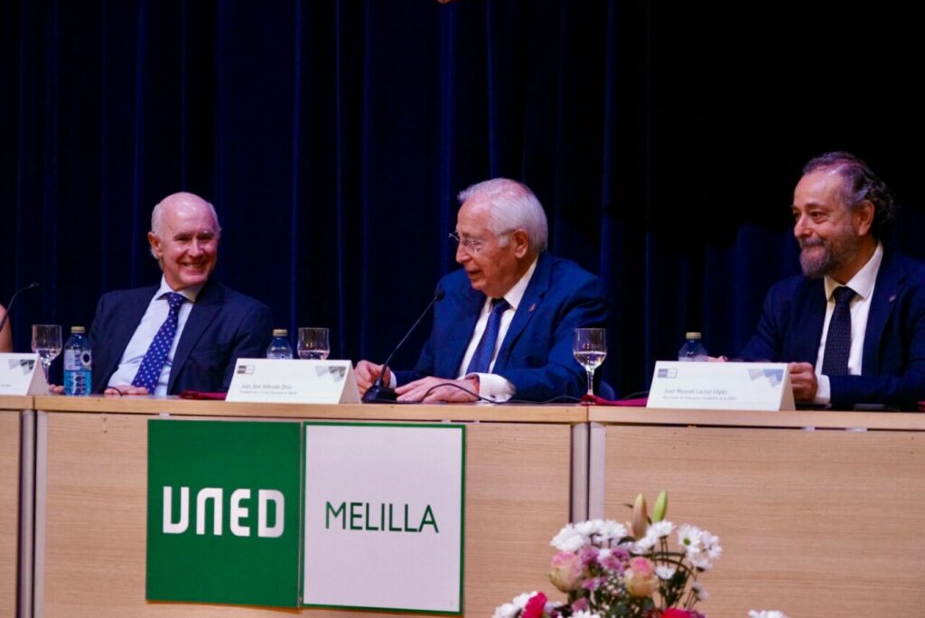 Panel of three men in suits seated at a conference table with microphones; UNED and MELILLA signs visible on the front panel and floral arrangement to the right.