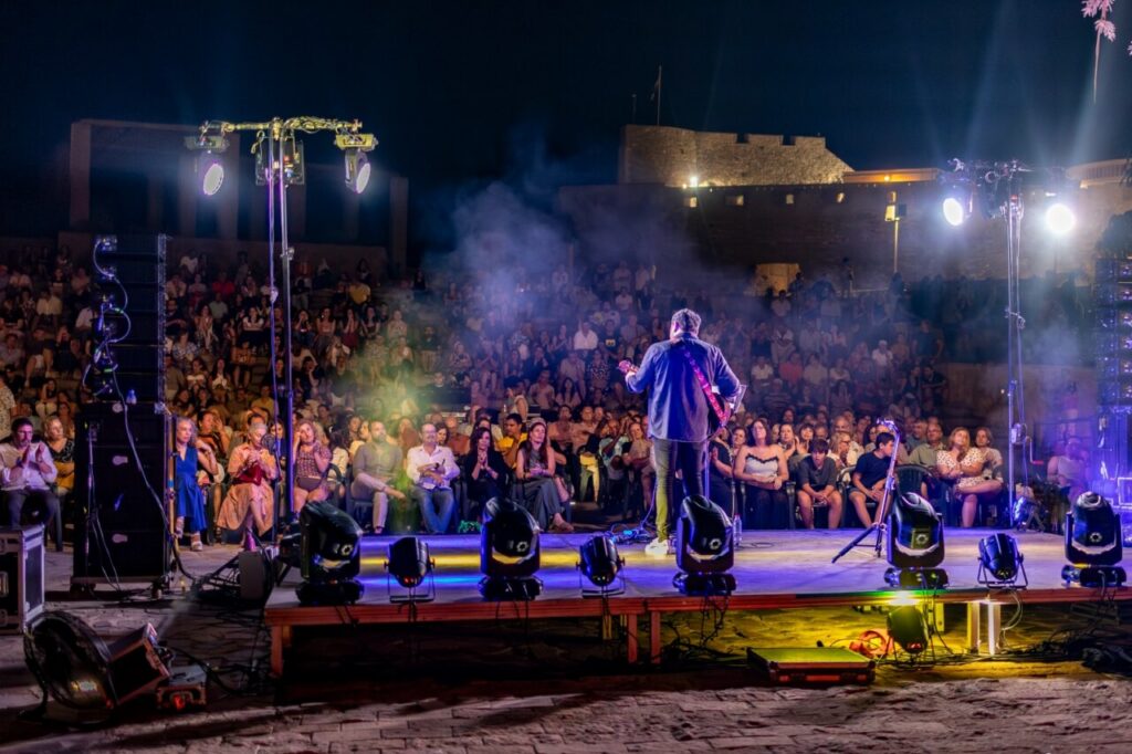 Musician with a guitar performing on a brightly lit stage to a packed outdoor audience at night, with fog and colorful lights.