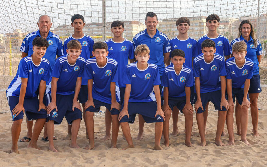 Group of young soccer players and two coaches posing for a team photo on a sandy beach behind a goal net, all in blue uniforms.