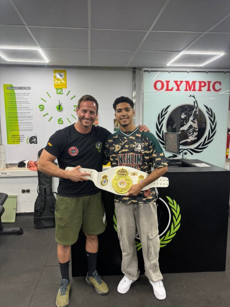 Two men stand in a gym, smiling as they hold a white championship belt with gold plates between them.