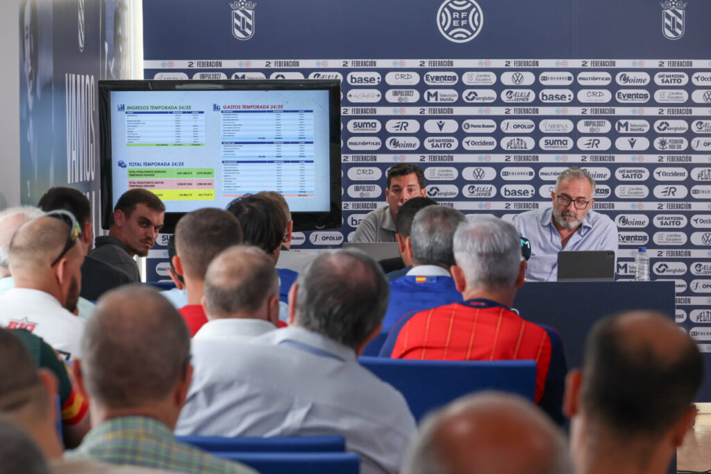 Panel of two men at a conference table presenting to an audience, with a large data screen beside them and a logoed backdrop behind them a press briefing setting.