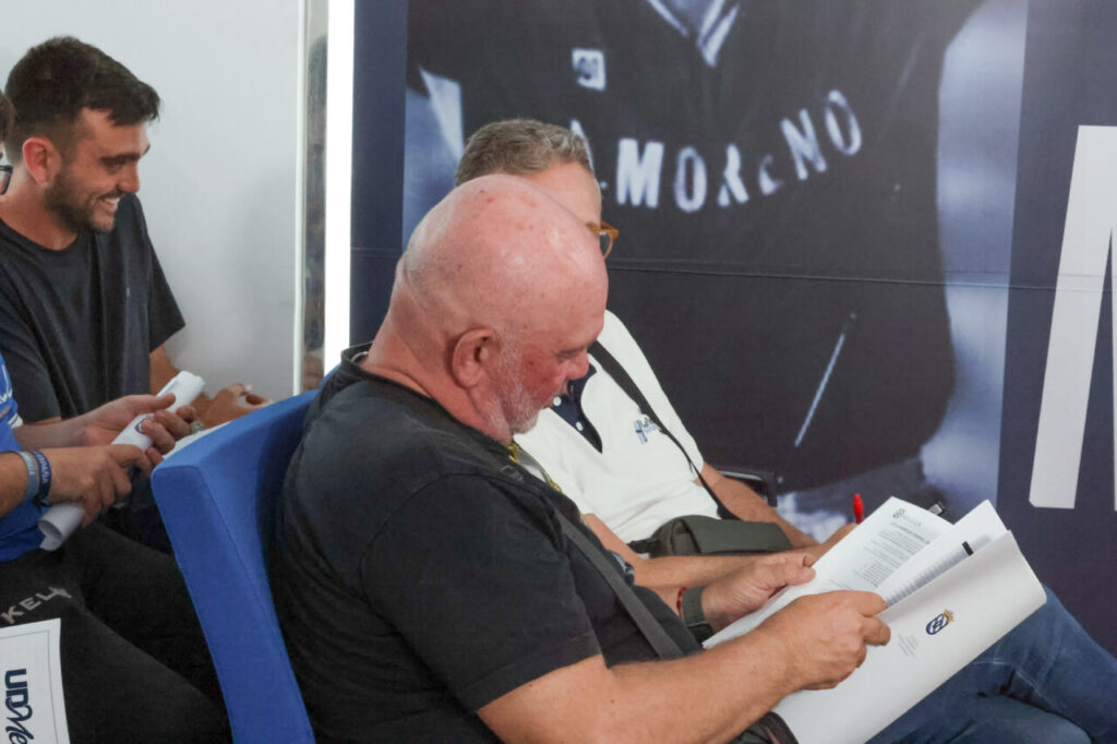 Bald man in a black T-shirt sits in a blue chair, reading a program at an event.